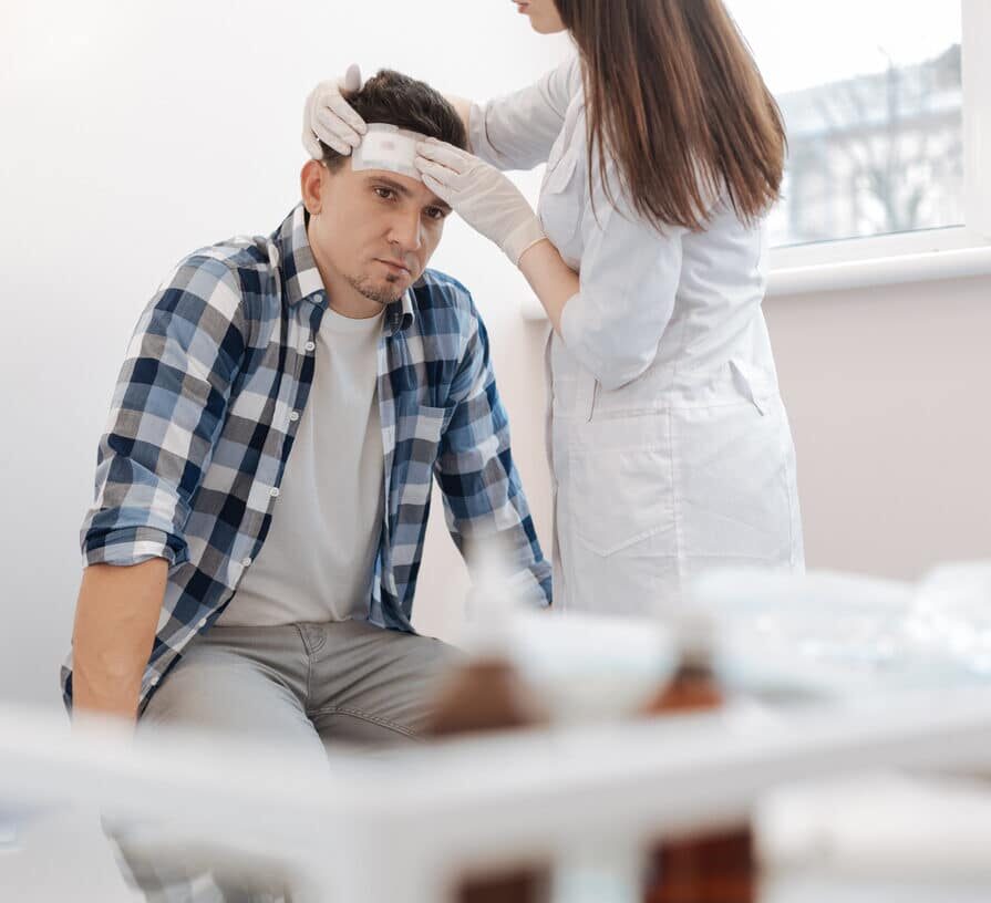 man sitting on the medical bed and receiving the first aid while having the forehead wound